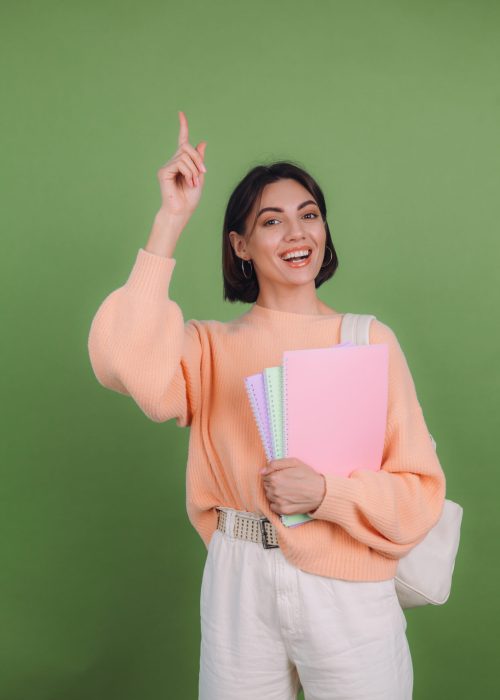 Young woman in casual peach sweater isolated on green olive color background with notebooks excited pointing finger up, idea came up, education high school concept